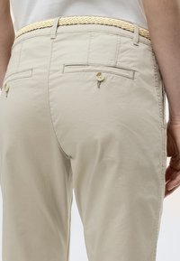 Εγγύς πλάνο μπεζ παντελονιού με πίσω τσέπες με κουμπιά και πλεκτή ζώνη ανοιχτού χρώματος, φορεμένο με λευκό πουκάμισο που είναι μέσα στην παντελόνα.