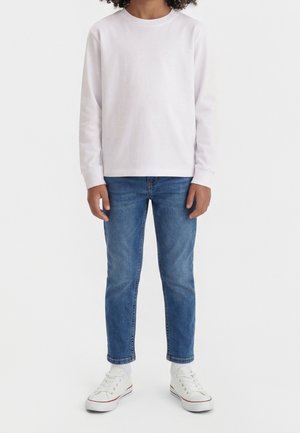 Barn iført hvid langærmet skjorte, blå jeans, hvide sokker og hvide sneakers stående mod en ensfarvet baggrund.