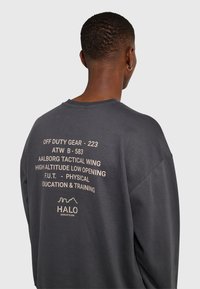 Persoon met kort haar en oorring in de vorm van een cirkel, van achteren gezien, draagt een donkergrijze trui met beige militaire tekst op de achterkant.