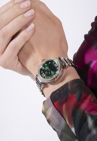 Roheline dialiga randme kell, millel on kuldsed aktsendid ja rooma numbrid, roostevabast terasest rihm ning dekoratiivne bezel, mis on kaunistatud kalliskividega.