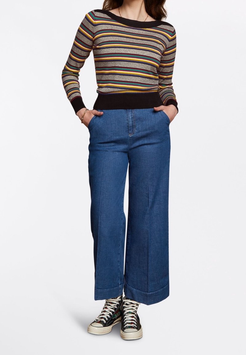 Gestreifter Langarmpullover mit einem schwarzen Saum, kombiniert mit weit geschnittenen blauen Jeans und schwarzen Sneakers mit weißen Sohlen und Schnürsenkeln.