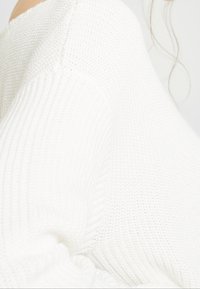 Maglione bianco lavorato a maglia con una texture a coste, caratterizzato da un ampio scollo e un design morbido. Il tessuto appare morbido e leggero.