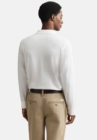 Pitkähihainen valkoinen poolopaita klassisella kauluksella, yhdistettynä beigenvärisiin housuihin, joissa on takataskut ja ruskea vyö. Sileä pinta.