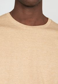 Beige kereknyakú póló finom bordázással a galléron és diszkrét textúrával, semleges háttér előtt bemutatva.