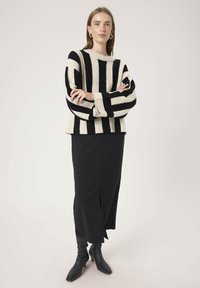 Frau in einem schwarz-beigen gestreiften Pullover mit Ballonärmeln, einem schwarzen Midikleid und schwarzen High Heel Stiefeln, die mit verschränkten Armen steht.
