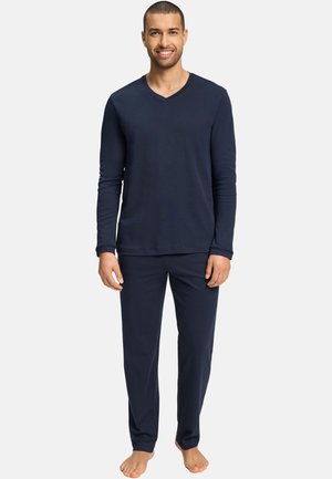 Homme debout pieds nus portant un haut à manches longues bleu marine et un pantalon assorti, souriant sur un fond neutre.
