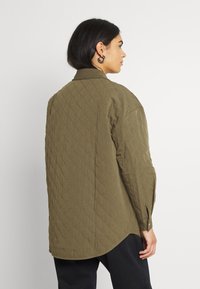 Jaqueta de camisa em verde oliva acolchoada, com colarinho, mangas longas com punhos botoados e uma bainha curva. Padrão de diamante texturizado por toda a peça.