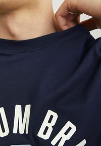 Tamno plava pamučna majica s okruglim izrezom, koja na prednjoj strani ima bijeli natpis. Tkanina izgleda mekano s glatkom teksturom.