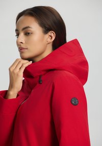 Veste imperméable rouge avec un col haut, une capuche ajustable et un patch logo noir sur la manche. Texture lisse et design minimaliste.