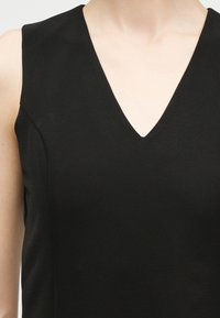 Μαύρο τοπ χωρίς μανίκια με λαιμόκοψη σε σχήμα V, φορεμένο από ένα άτομο, που δείχνει το πάνω μέρος του στήθους και τους ώμους, σε λευκό φόντο.