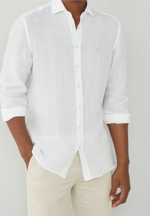 Homme portant une chemise blanche à manches longues retroussées et un pantalon beige, debout avec une main dans la poche.