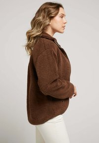 Veste en polaire marron avec fermeture éclair, col montant, poches latérales et coupe légèrement oversize. Tissu texturé au toucher doux.
