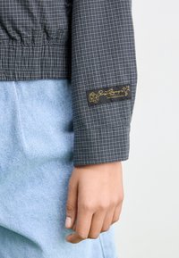 Svart og grå rutete skjorte med elastiske mansjetter og en liten hvit etikett med gulltekst. Blå denimbukser delvis synlige.