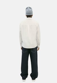 Camicia bianca a maniche lunghe con yoke posteriore, abbinata a jeans a gamba larga blu scuro. Il modello indossa un cappellino lavorato a maglia grigio chiaro.