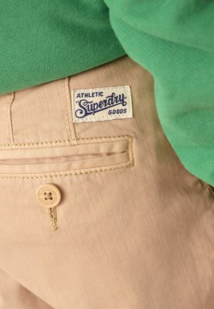 Béžové bavlnené nohavice s prešitým vreckom, so smotanovou etikou s modrým textom a hnedým gombíkom s vyrytým nápisom "Superdry". Zelený kus oblečenia nad nimi.