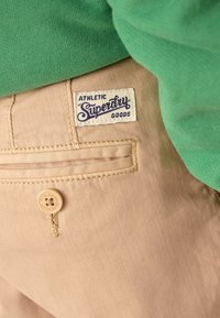 Pantalon en coton beige avec une poche arrière ornée d'une étiquette « Superdry » et un bouton marron clair. Tissu vert partiellement visible au-dessus du pantalon.