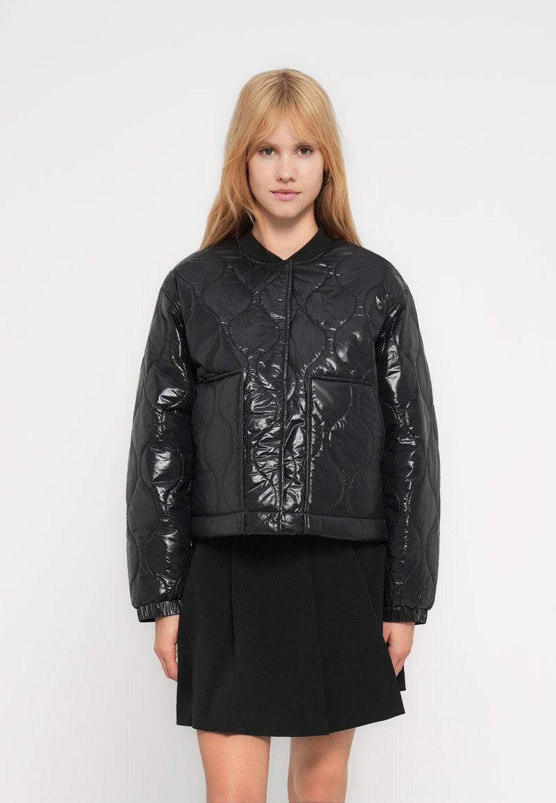 Νέα γυναίκα με ίσια ξανθά μαλλιά, φορώντας μια γυαλιστερή, καπιτονέ μαύρη ζακέτα και μια μαύρη πλισέ φούστα, σε λευκό φόντο.