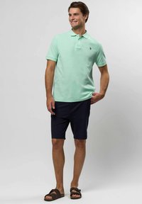 Mężczyzna w miętowej koszulce polo, granatowych szortach i brązowych sandałach, stoi i uśmiecha się na tle jednolitego szarego tła.