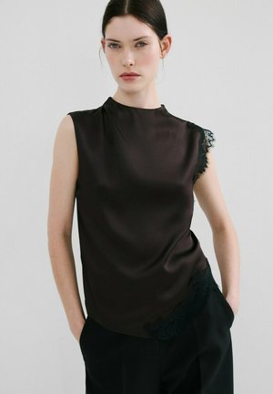 Mujer de cabello oscuro que lleva una blusa sin mangas de satén negro con detalles de encaje, de pie con las manos en los bolsillos frente a un fondo liso.