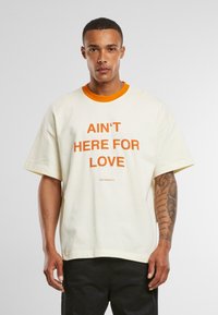 T-shirt crème à manches courtes avec bord orange et texte en gras "JE NE SUIS PAS LÀ POUR L'AMOUR", offrant une coupe décontractée et une texture douce.