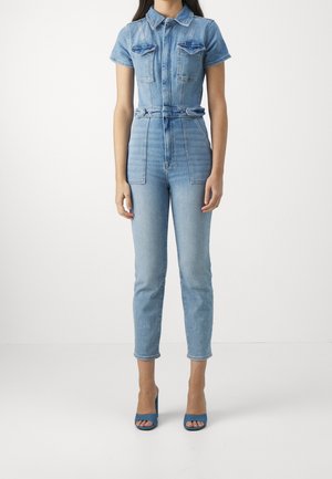 Kvinde iført en lyseblå denim-jumpsuit med korte ærmer, brystlommer, et figursyet bælte i taljen, korte ben og blå åbne sko med hæl.