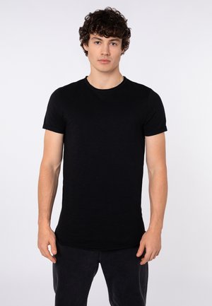 Young man with curly hair wearing a plain black fitted short-sleeve t-shirt and dark pants, facing forward against a white background.