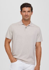 Polo de color beige claro hecho de una tela suave y texturizada. Presenta un cuello, mangas cortas y dos botones oscuros en el escote.