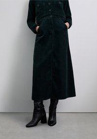 Jupe en velours côtelé vert foncé avec une longue silhouette évasée en A, dotée de poches latérales et d'une fermeture à boutons, portée avec des bottines noires.