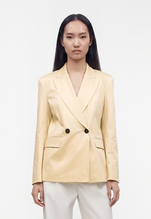 Vrouw met lang zwart haar draagt een beige double-breasted blazer met zwarte knopen en witte broek tegen een effen lichte achtergrond.