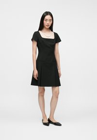 Μαύρο φόρεμα με κοντά μανίκια, τετράγωνη λαιμόκοψη, εφαρμοστό μπούστο και φουσκωτή φούστα; λεία ύφασμα; μοντέλο φοράει μαύρες μπαλαρίνες.