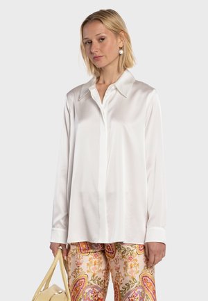 Kvinde iført en hvid silkeagtig bluse og farverige mønstrede bukser, der holder en beige håndtaske mod en ensfarvet baggrund.