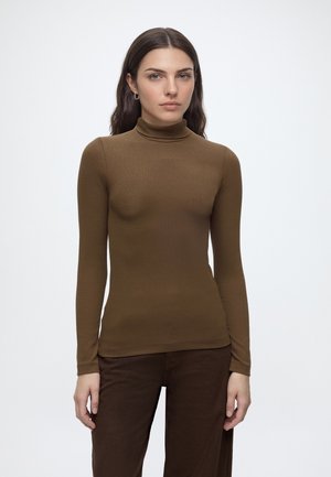 Vrouw met lang donker haar, die een aansluitende bruine coltrui en donkerbruine broek draagt, staande tegen een effen lichte achtergrond.