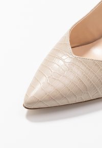 Chaussure beige à bout pointu avec un motif crocodile texturé. Comprend une doublure intérieure lisse et une semelle en cuir.