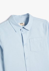 Camicia a maniche corte di cotone azzurro chiaro con colletto a punte, chiusura frontale con bottoni e una singola tasca sul petto con contorno cucito.