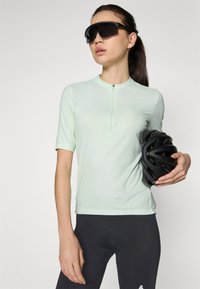 Mintgroene fietsshirt met korte mouwen en een ritssluiting bij de hals, gemaakt van een zachte stof, gecombineerd met zwarte fietsshorts en een zwarte helm.
