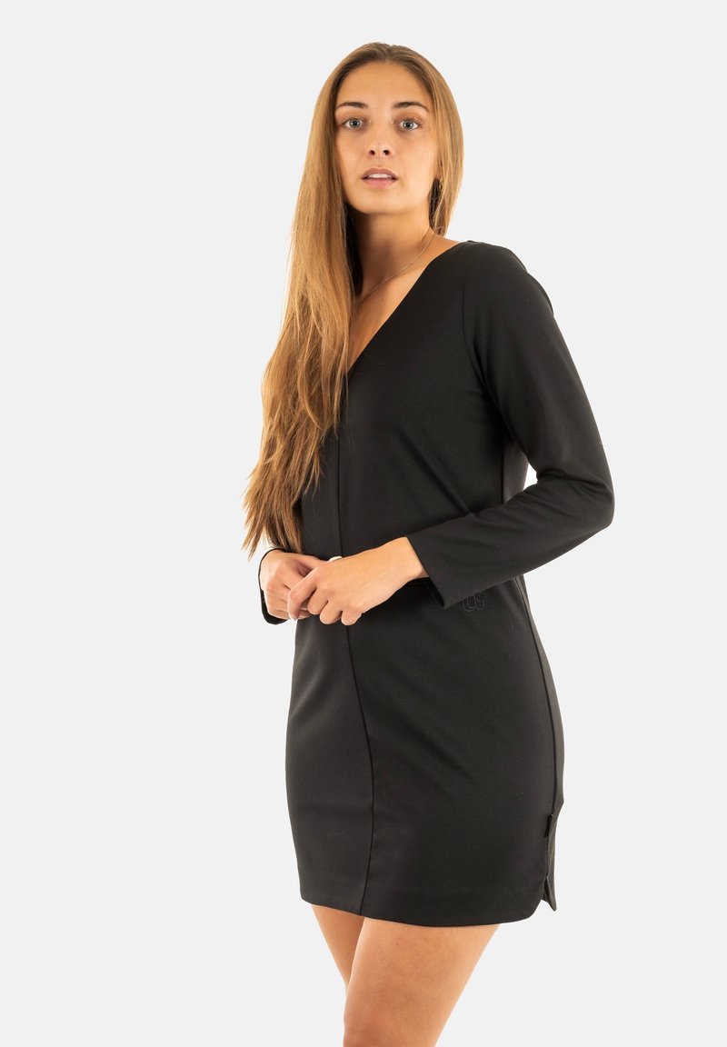 Frau mit langen Haaren, die ein schwarzes, langärmliges V-Ausschnitt-Kleid trägt, steht mit gefalteten Händen vor einem schlichten hellen Hintergrund.