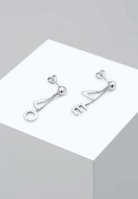Pendientes de plata con formas circulares y angulares, conectados por cadenas, con acentos esféricos. Expuestos en un pedestal minimalista blanco.
