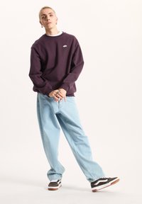Fioletowy sweter z okrągłym dekoltem z małym logo, w zestawieniu z jasnoniebieskimi spodniami typu wide-leg i czarno-białymi sneakerami z białymi akcentami.