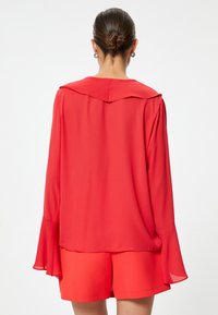 Czerwona bluzka z szerokim kołnierzem i rozkloszowanymi długimi rękawami, wykonana z gładkiego materiału. Stylizowana z pasującymi czerwonymi szortami widocznymi na dole.