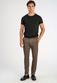 Mand står iført en tætsiddende sort t-shirt, brune slim pants og sorte læderslippers, med den ene hånd i lommen.