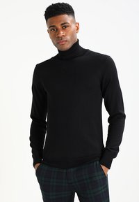 Schwarzer Rollkragenpullover aus strukturiertem Strick. Figurnahes Design mit langen Ärmeln, kombiniert mit grün-blau karierten Hosen.