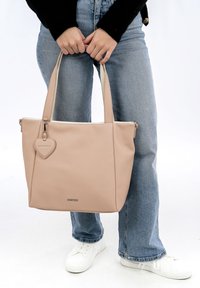 Beige kožená taška přes rameno se strukturovaným designem, vybavená dvěma uchy a přívěskem ve tvaru srdce, na světlém pozadí.