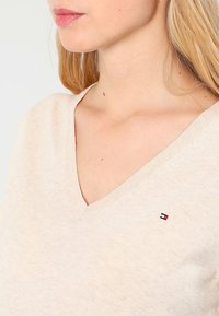 Kobieta z długimi blond włosami, ubrana w beżowy sweter w serek z małym prostokątnym logo w kolorach czerwonym, białym i niebieskim na piersi.