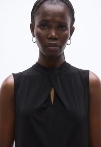 Fekete ujjatlan blúz, csomós részlettel a nyakkivágásnál és egy finom kulcslyuk alakú kivágással, sima, könnyű anyagból készült.