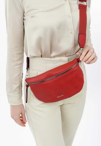 Borsa a tracolla in pelle rossa con forma curva, dotata di una zip in argento, una tracolla in tessuto e un piccolo dettaglio del logo sul fronte.