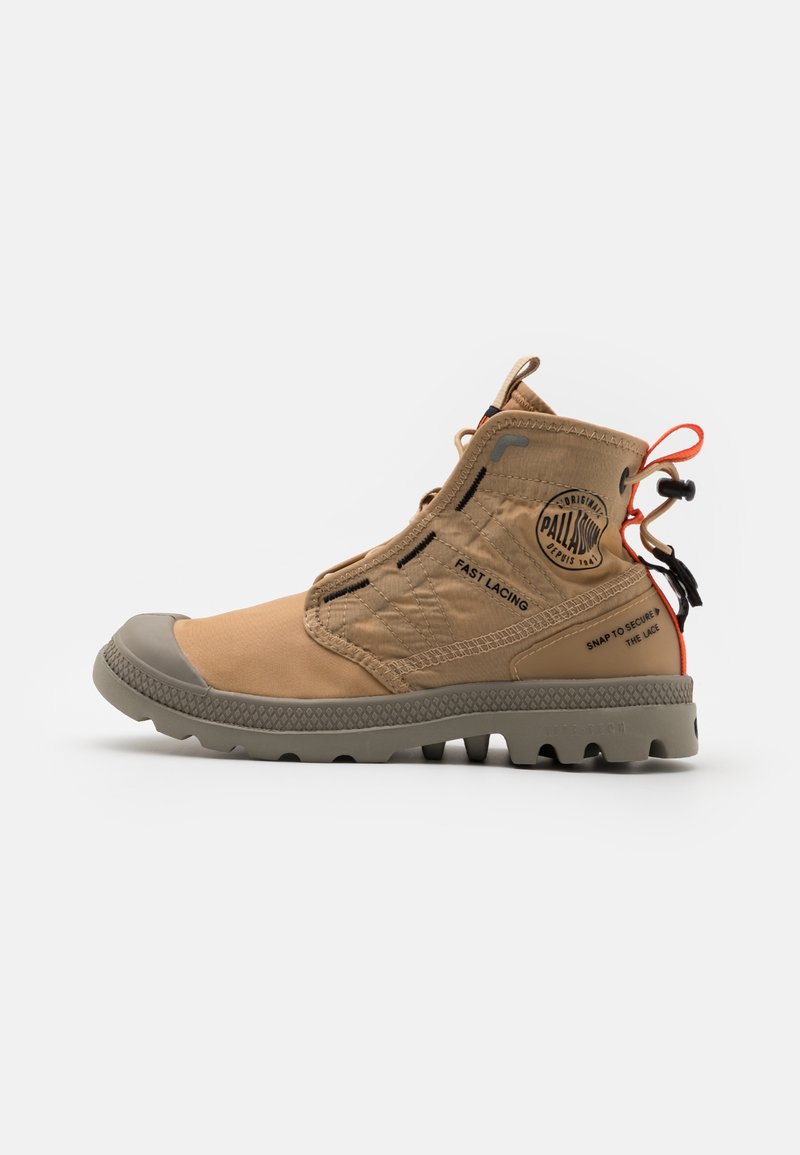 Botte haute de couleur beige avec une semelle grise robuste, un système de laçage rapide, des tirettes noires et orange, et le logo de la marque Palladium sur le côté.