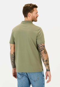 Olijfgroene polo met kraag, korte mouwen en zijsplitten, gedragen met lichtblauwe spijkerbroek. Tatoeages zichtbaar op de arm.