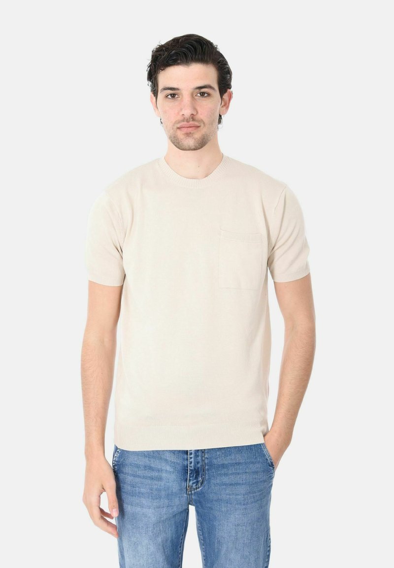 Maglietta a maniche corte in maglia beige con collo a giro e taschino sul petto, abbinata a jeans azzurri chiari. Texture morbida e design aderente.