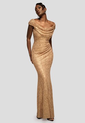 Mujer elegante posa con un vestido largo hasta el suelo, dorado y cubierto de lentejuelas, con un escote drapeado que deja los hombros al descubierto, sobre un fondo sencillo.