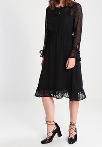 Černé šaty po kolena s puntíkovým vzorem, průhlednými dlouhými rukávy, uvazováním na krku, elastickým pasem a volánkovým lemem. V kombinaci s černými lodičkami.